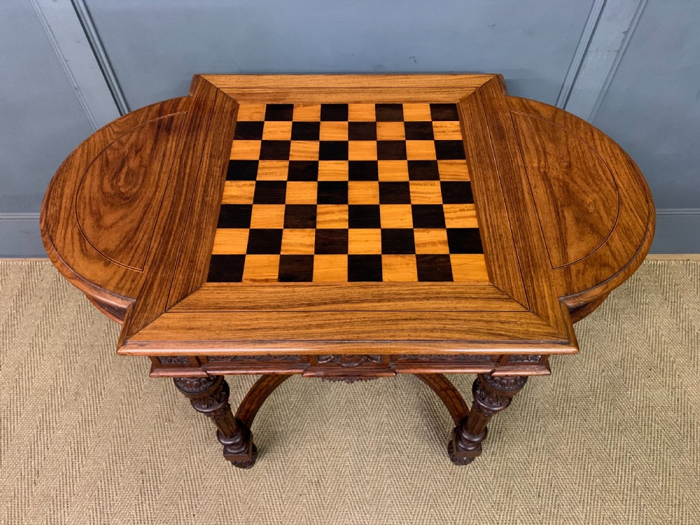 An Anglo-Indian rosewood games table featuring an inlaid square chessboard top, drop leaves, a carved frieze, and turned legs connected by a curved stretcher. View IV
