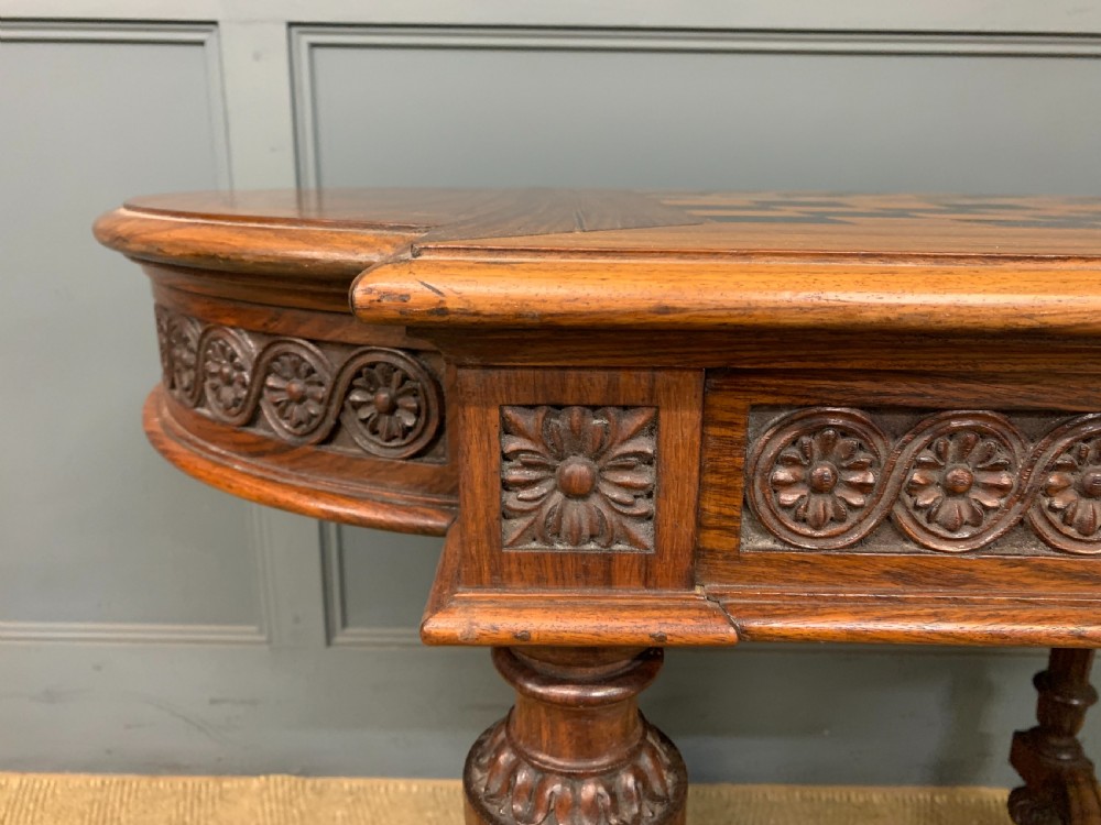 An Anglo-Indian rosewood games table featuring an inlaid square chessboard top, drop leaves, a carved frieze, and turned legs connected by a curved stretcher. View VI
