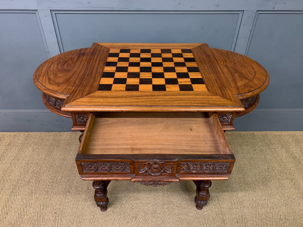 An Anglo-Indian rosewood games table featuring an inlaid square chessboard top, drop leaves, a carved frieze, and turned legs connected by a curved stretcher. View X
