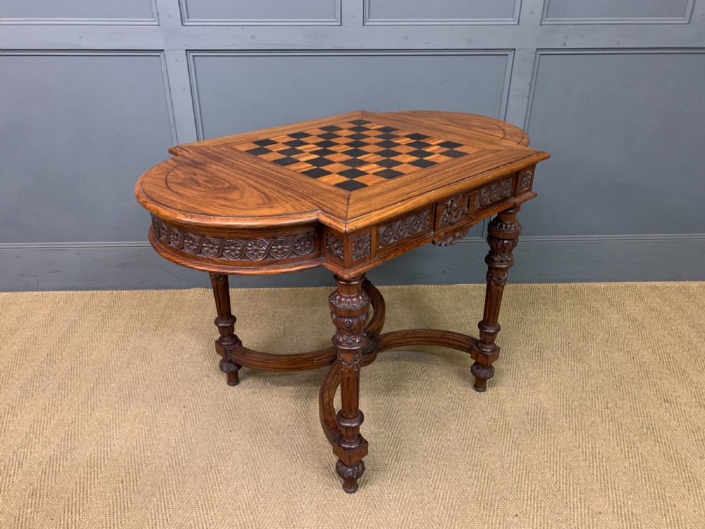 An Anglo-Indian rosewood games table featuring an inlaid square chessboard top, drop leaves, a carved frieze, and turned legs connected by a curved stretcher. View XI