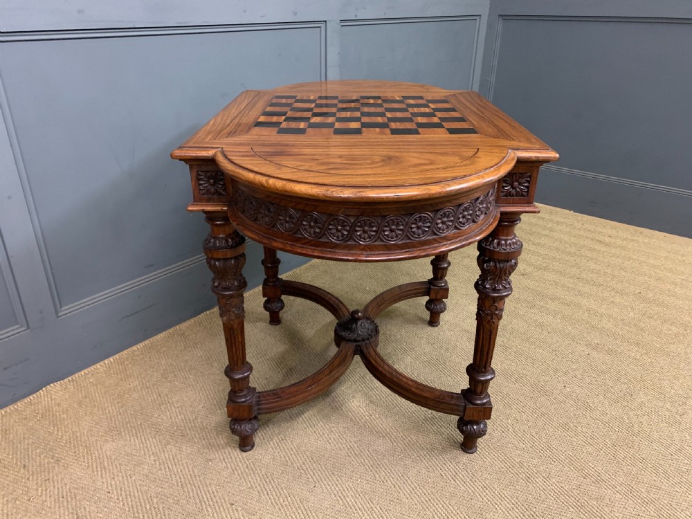 An Anglo-Indian rosewood games table featuring an inlaid square chessboard top, drop leaves, a carved frieze, and turned legs connected by a curved stretcher. View XII