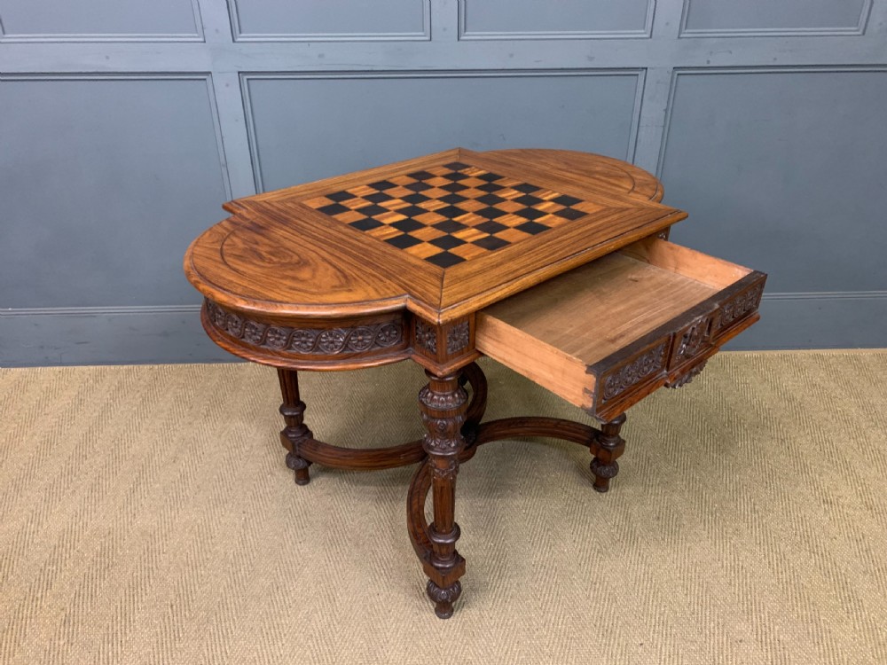 An Anglo-Indian rosewood games table featuring an inlaid square chessboard top, drop leaves, a carved frieze, and turned legs connected by a curved stretcher. View XVI