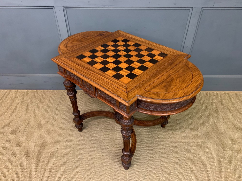 An Anglo-Indian rosewood games table featuring an inlaid square chessboard top, drop leaves, a carved frieze, and turned legs connected by a curved stretcher. View XVII