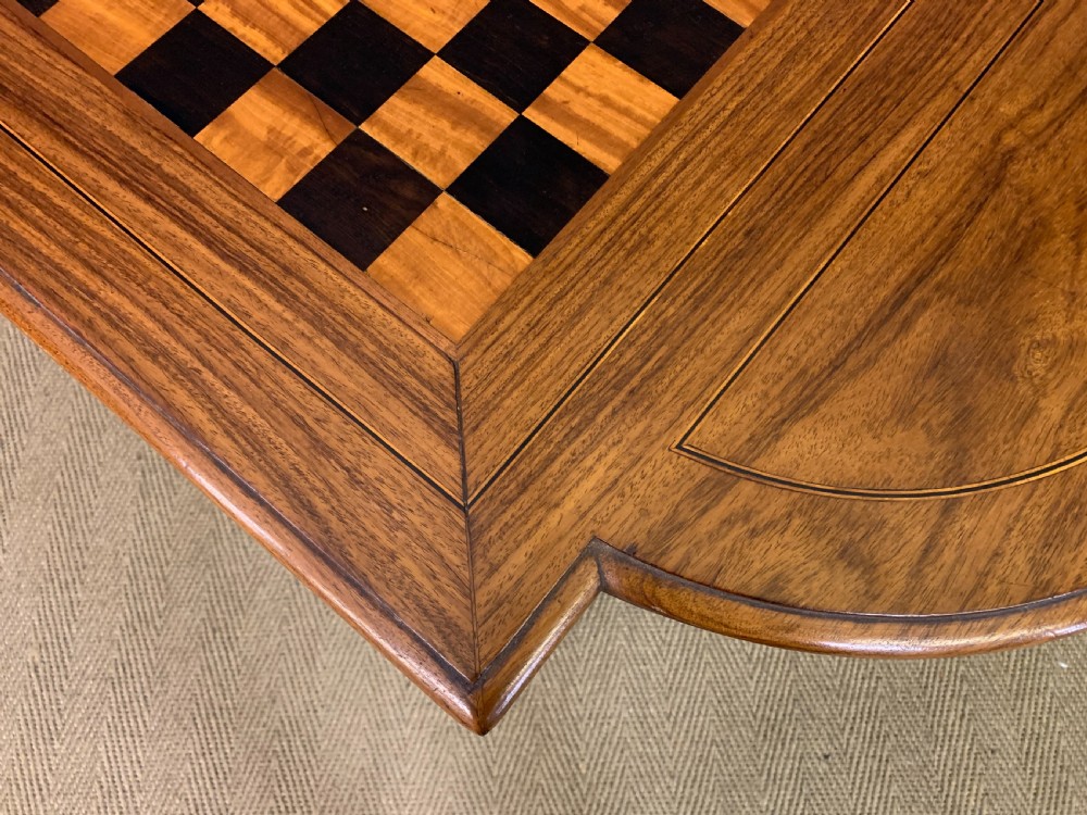 An Anglo-Indian rosewood games table featuring an inlaid square chessboard top, drop leaves, a carved frieze, and turned legs connected by a curved stretcher. View XVIII
