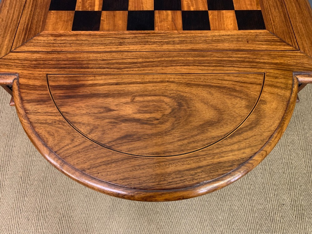 An Anglo-Indian rosewood games table featuring an inlaid square chessboard top, drop leaves, a carved frieze, and turned legs connected by a curved stretcher. View XIX