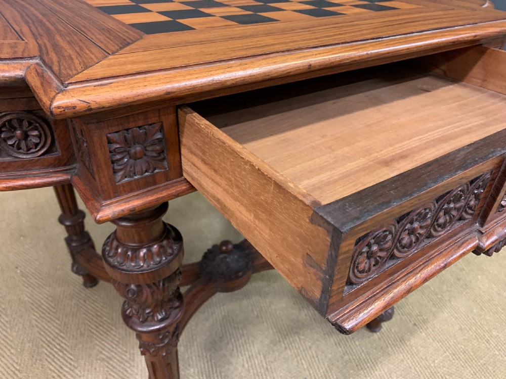 An Anglo-Indian rosewood games table featuring an inlaid square chessboard top, drop leaves, a carved frieze, and turned legs connected by a curved stretcher. View XX