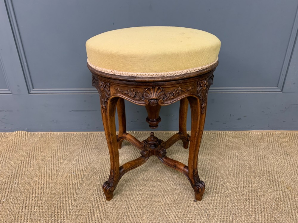 A French carved walnut adjustable piano stool with cabriole legs, carved floral apron, X-shaped stretcher, and a round light yellow upholstered seat.