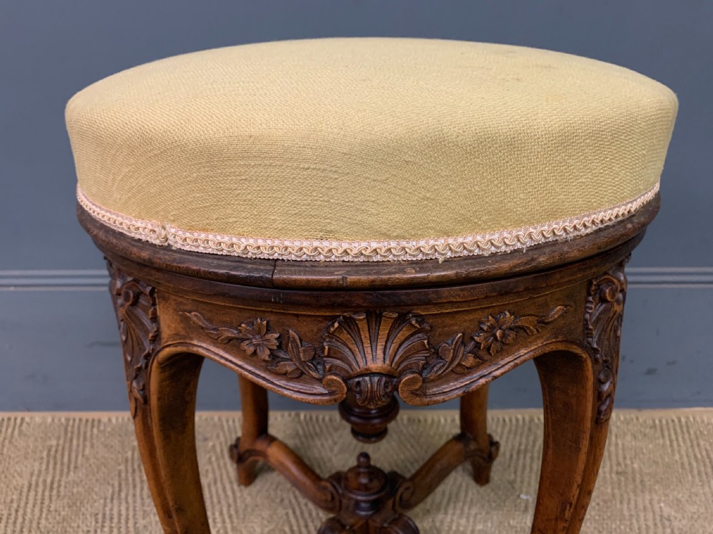 A French carved walnut adjustable piano stool with cabriole legs, carved floral apron, X-shaped stretcher, and a round light yellow upholstered seat. View II