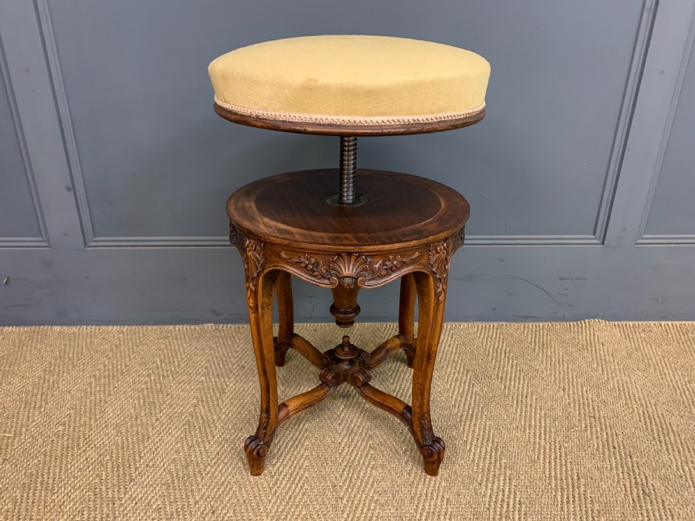 A French carved walnut adjustable piano stool with cabriole legs, carved floral apron, X-shaped stretcher, and a round light yellow upholstered seat. View IX