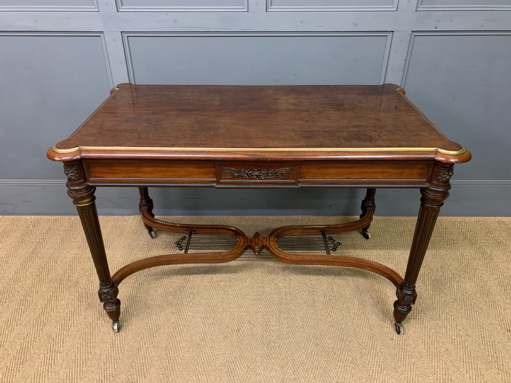 A rectangular French mahogany library table featuring a polished top, frieze drawer with carved detail, brass trim, turned fluted legs, and a lower carved stretcher on brass castors. View II