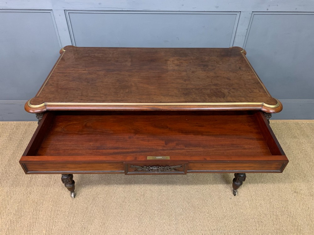 A rectangular French mahogany library table featuring a polished top, frieze drawer with carved detail, brass trim, turned fluted legs, and a lower carved stretcher on brass castors. View XI