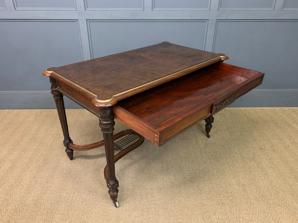 A rectangular French mahogany library table featuring a polished top, frieze drawer with carved detail, brass trim, turned fluted legs, and a lower carved stretcher on brass castors. View XIX