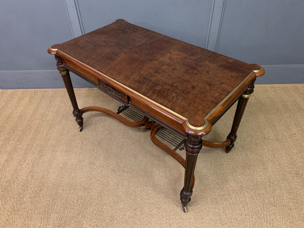 A rectangular French mahogany library table featuring a polished top, frieze drawer with carved detail, brass trim, turned fluted legs, and a lower carved stretcher on brass castors. View XX