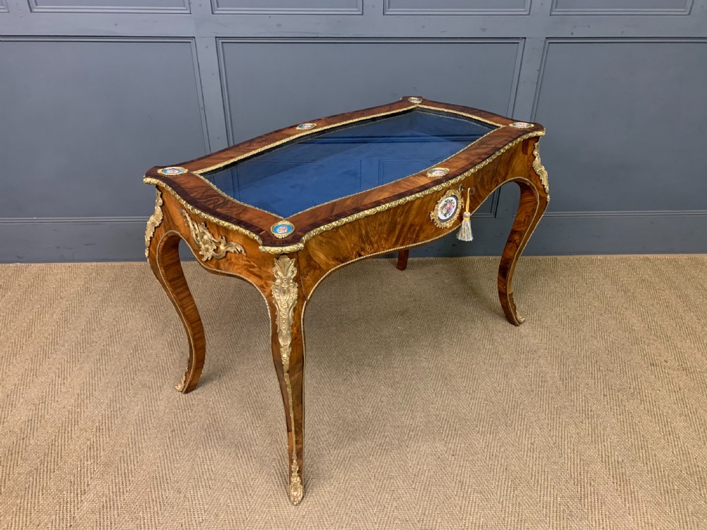 A 19th-century French Louis XV style bijouterie table with a glazed display top, curved cabriole legs, gilt bronze mounts, porcelain plaques, and a blue velvet interior.