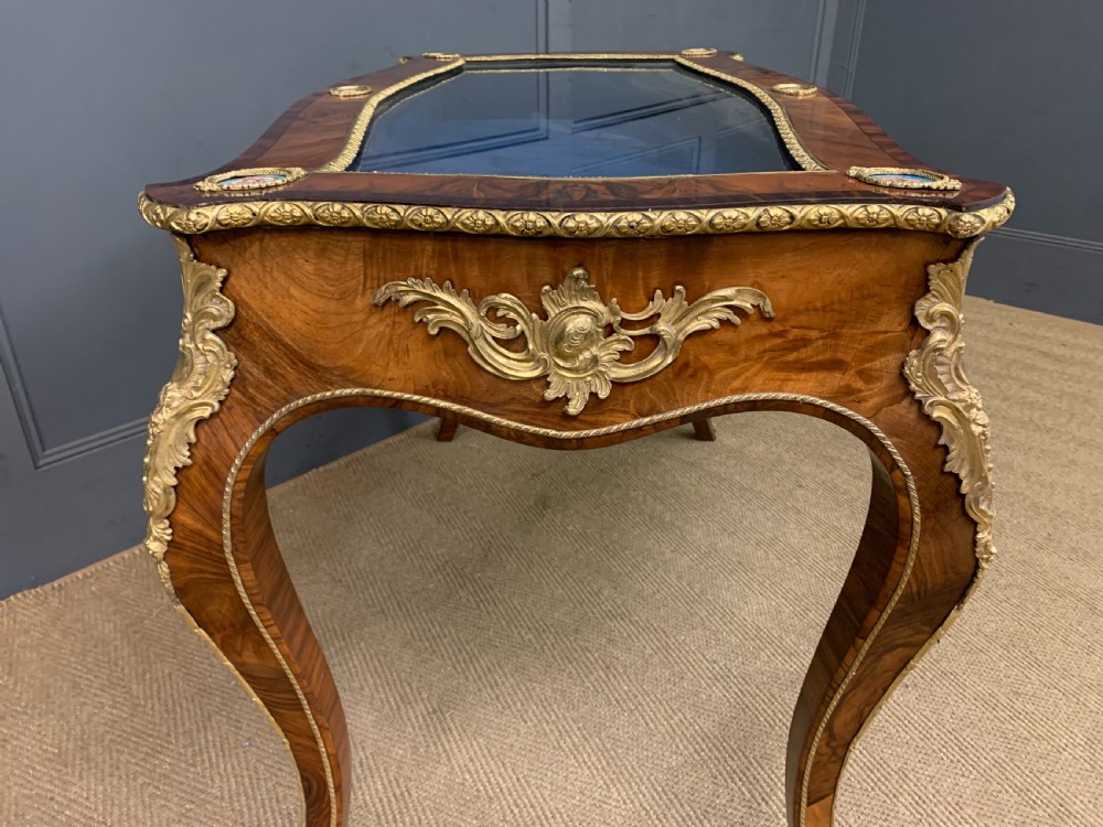 A 19th-century French Louis XV style bijouterie table with a glazed display top, curved cabriole legs, gilt bronze mounts, porcelain plaques, and a blue velvet interior. View X