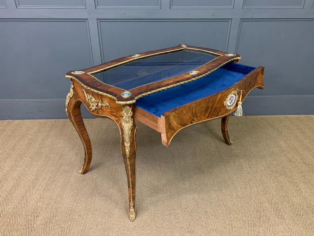 A 19th-century French Louis XV style bijouterie table with a glazed display top, curved cabriole legs, gilt bronze mounts, porcelain plaques, and a blue velvet interior. View XV