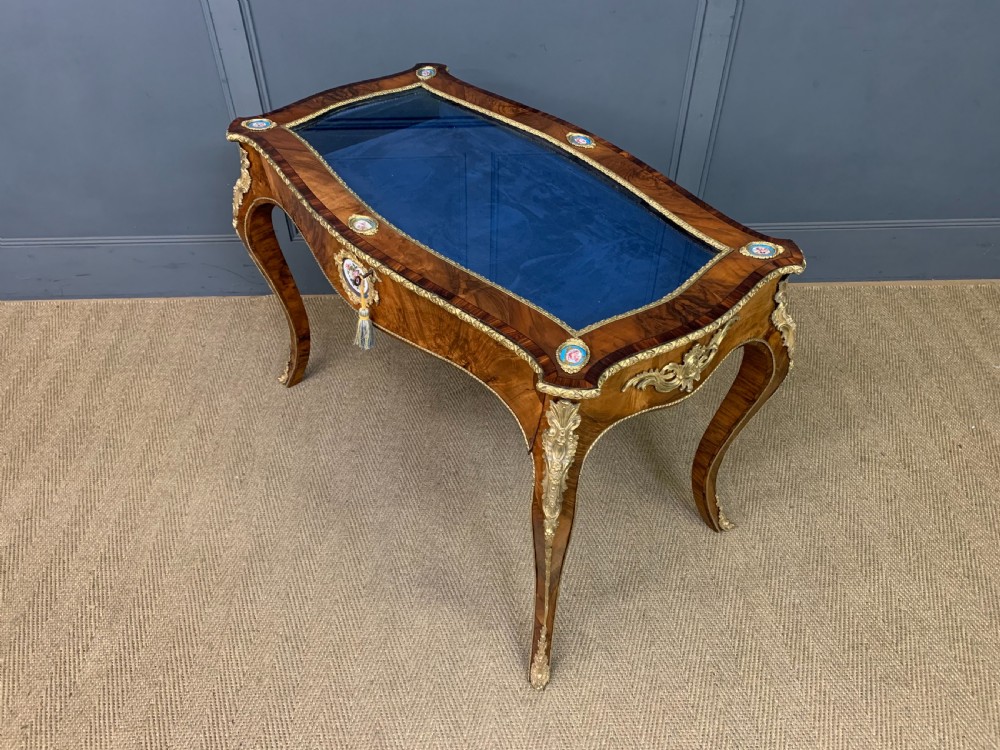 A 19th-century French Louis XV style bijouterie table with a glazed display top, curved cabriole legs, gilt bronze mounts, porcelain plaques, and a blue velvet interior. View XVI