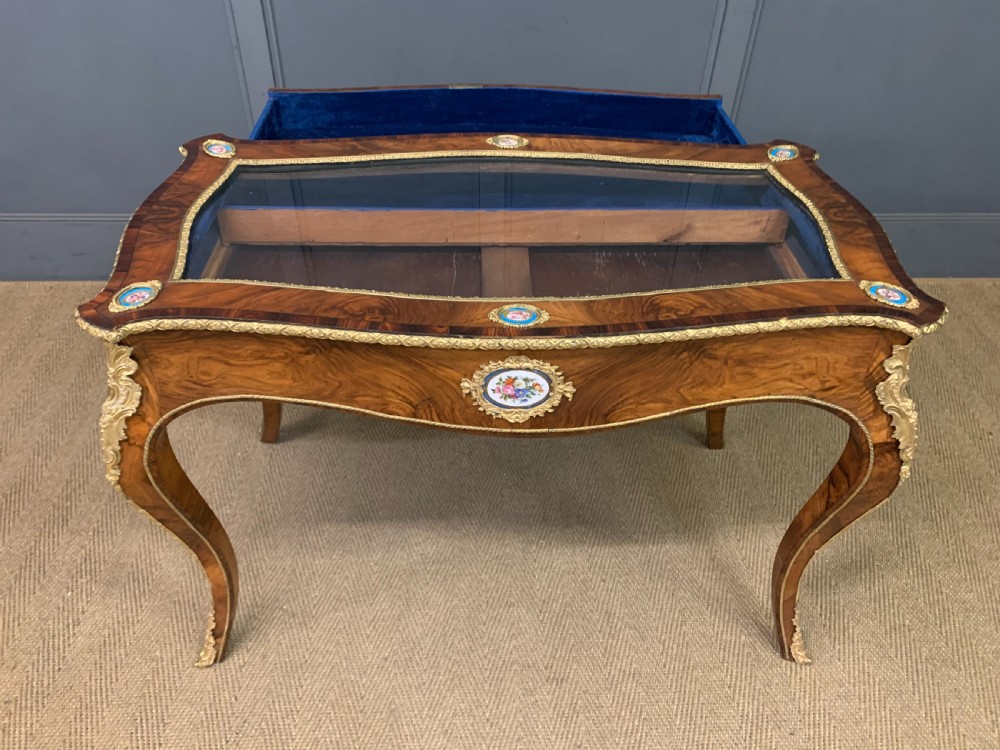 A 19th-century French Louis XV style bijouterie table with a glazed display top, curved cabriole legs, gilt bronze mounts, porcelain plaques, and a blue velvet interior. View XVIII