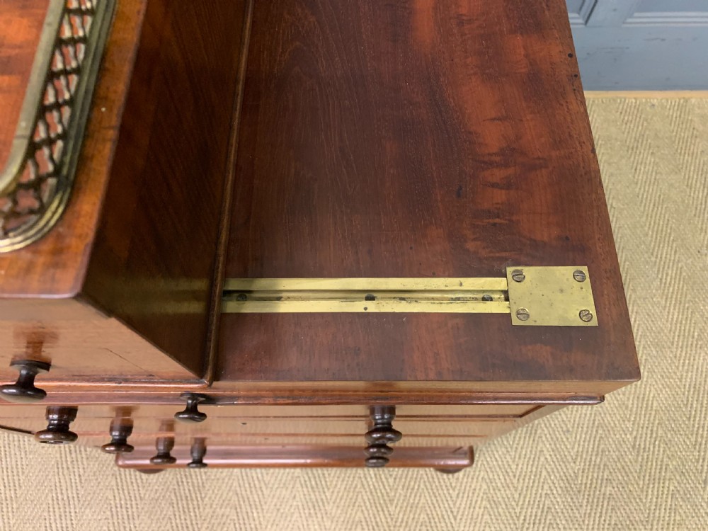 A George IV mahogany Davenport desk featuring a slanted brown leather writing surface, brass gallery rail, four side drawers with turned knobs, and bun feet. View XXX