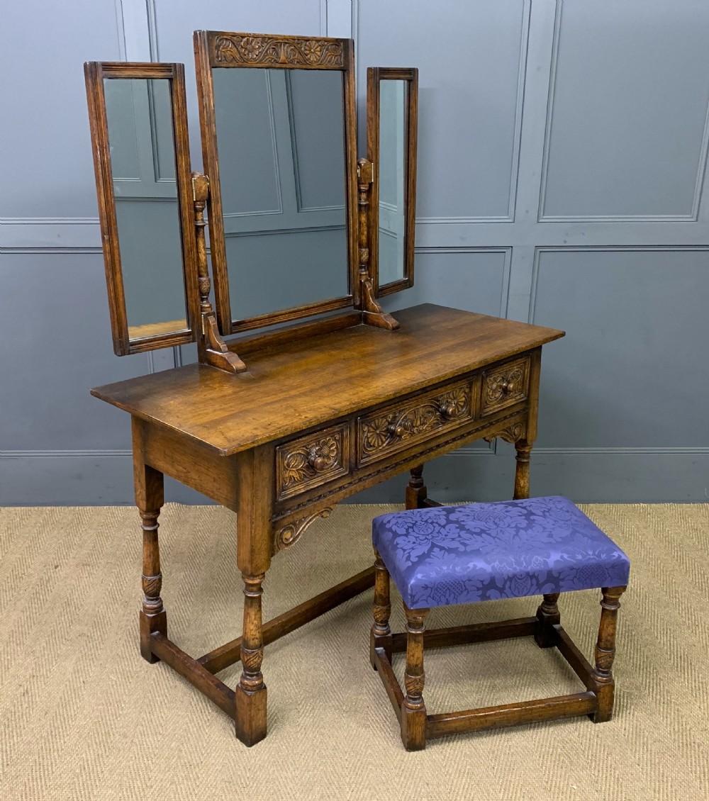 A carved oak dressing table featuring a central tri-fold mirror, three carved drawers, turned legs, and an accompanying stool upholstered in purple patterned fabric.