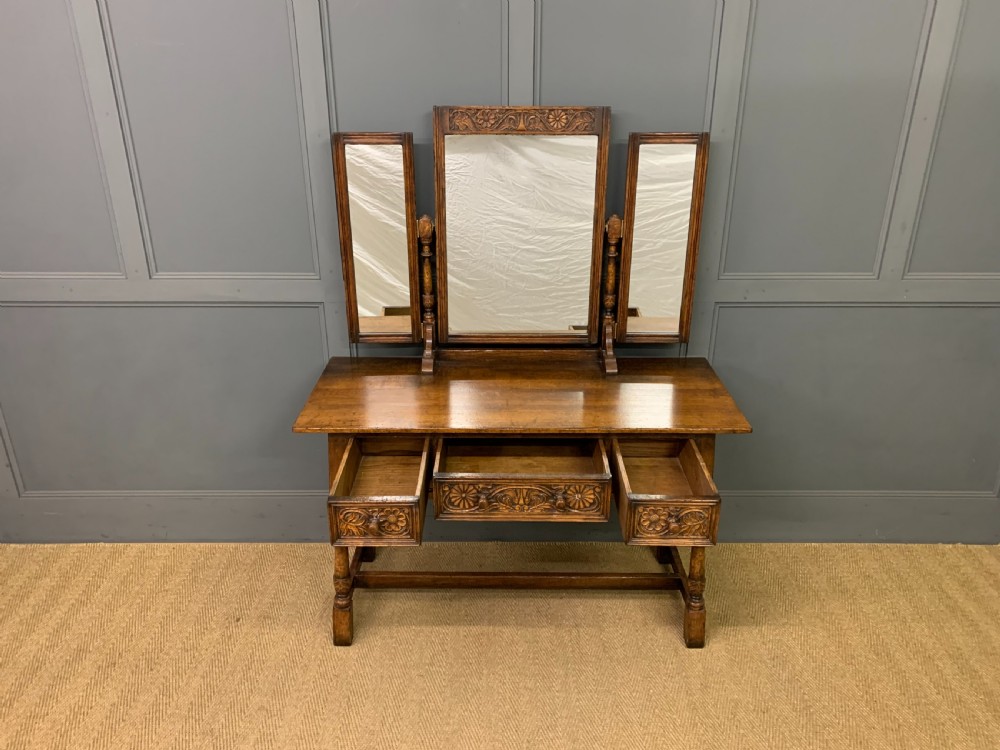 A carved oak dressing table featuring a central tri-fold mirror, three carved drawers, turned legs, and an accompanying stool upholstered in purple patterned fabric. View V
