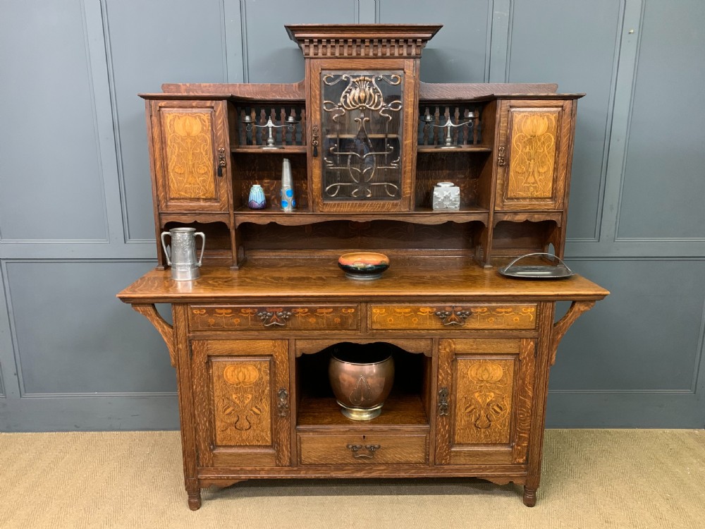 An Arts and Crafts oak sideboard featuring detailed inlaid floral marquetry panels, a central leaded glass upper cabinet, and decorative copper hardware.