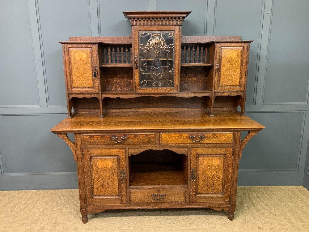 An Arts and Crafts oak sideboard featuring detailed inlaid floral marquetry panels, a central leaded glass upper cabinet, and decorative copper hardware. View III