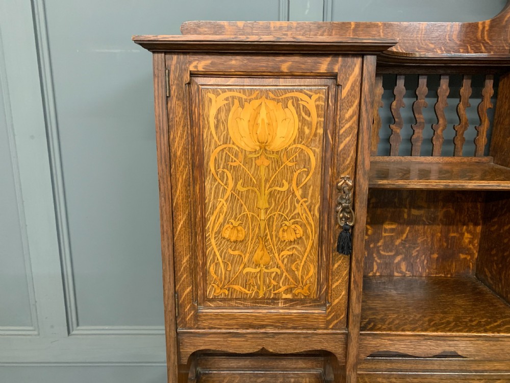 An Arts and Crafts oak sideboard featuring detailed inlaid floral marquetry panels, a central leaded glass upper cabinet, and decorative copper hardware. View IV