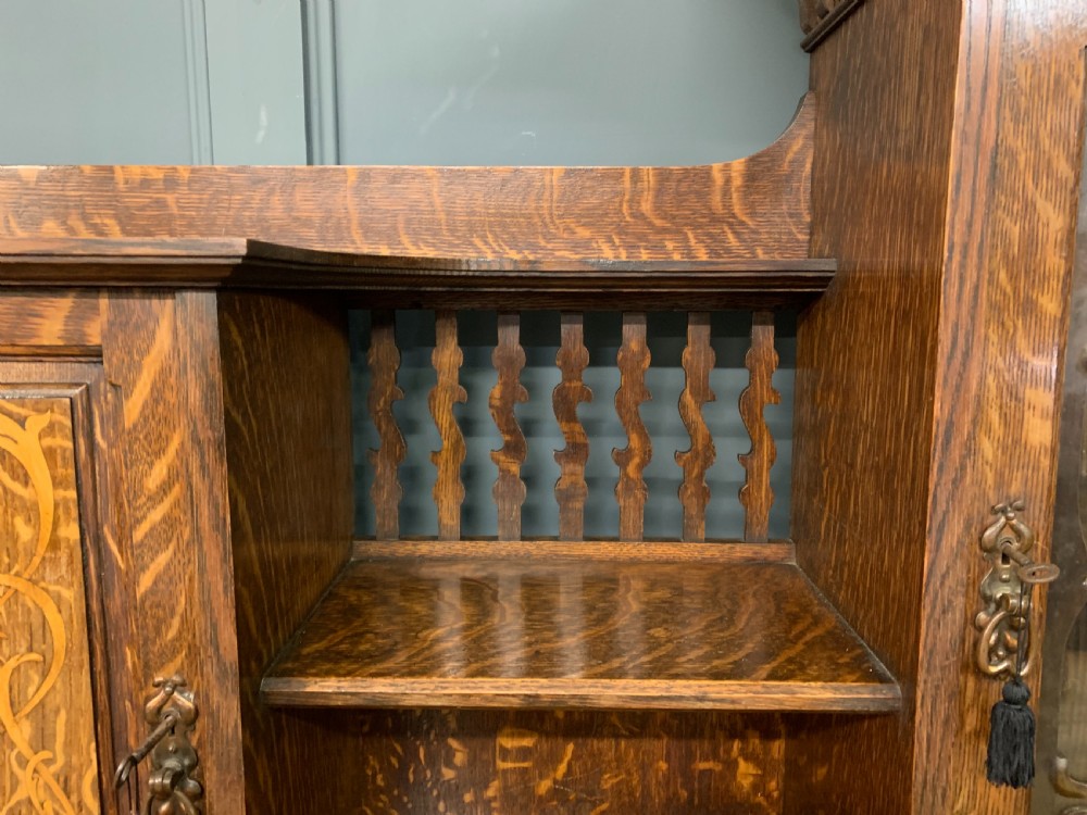 An Arts and Crafts oak sideboard featuring detailed inlaid floral marquetry panels, a central leaded glass upper cabinet, and decorative copper hardware. View V
