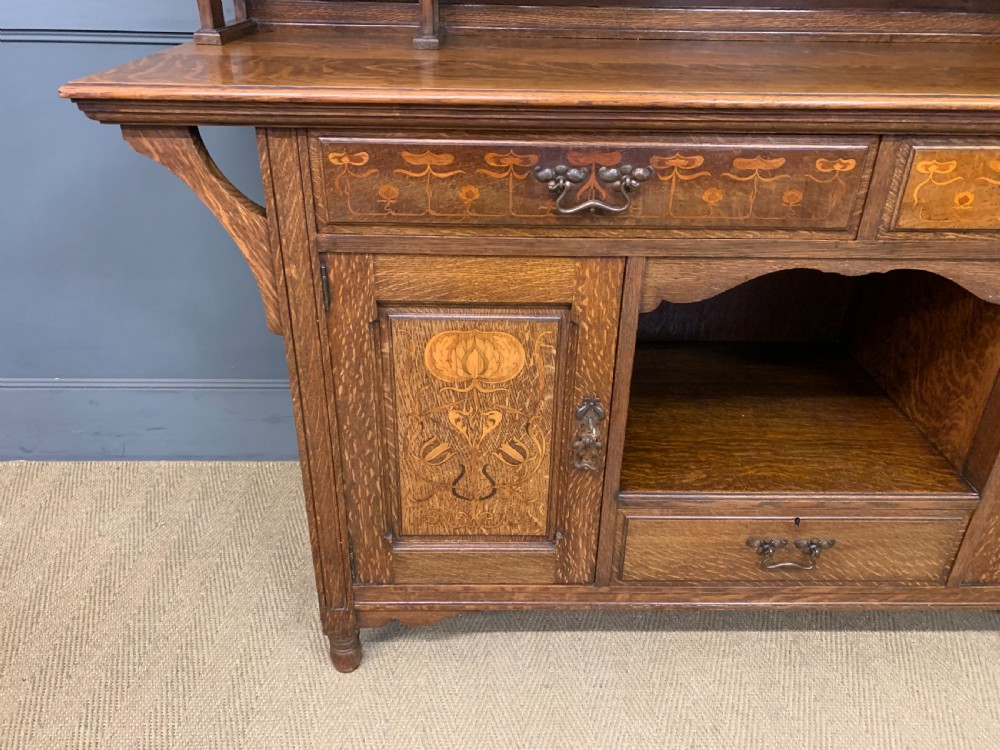An Arts and Crafts oak sideboard featuring detailed inlaid floral marquetry panels, a central leaded glass upper cabinet, and decorative copper hardware. View X