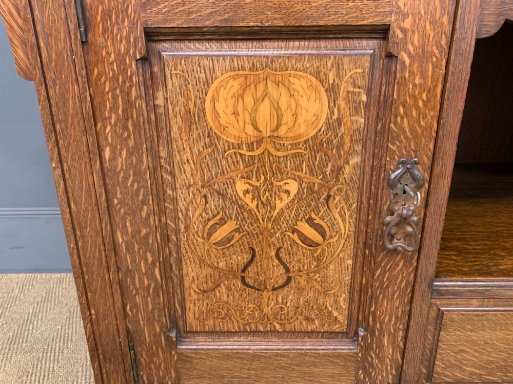 An Arts and Crafts oak sideboard featuring detailed inlaid floral marquetry panels, a central leaded glass upper cabinet, and decorative copper hardware. View XII
