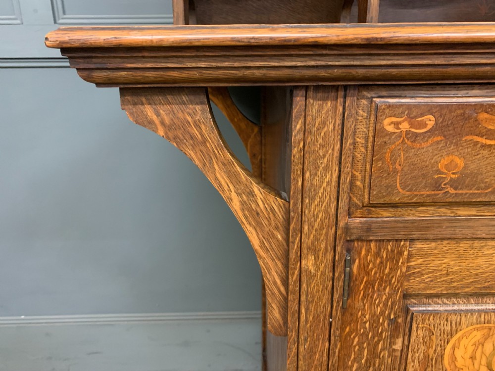 An Arts and Crafts oak sideboard featuring detailed inlaid floral marquetry panels, a central leaded glass upper cabinet, and decorative copper hardware. View XIV