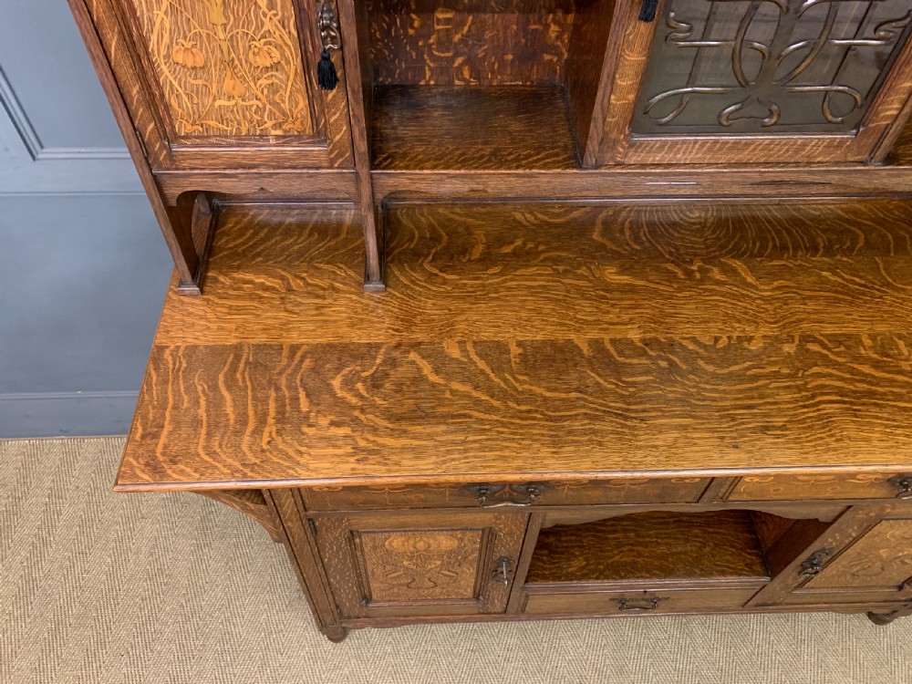An Arts and Crafts oak sideboard featuring detailed inlaid floral marquetry panels, a central leaded glass upper cabinet, and decorative copper hardware. View XVI