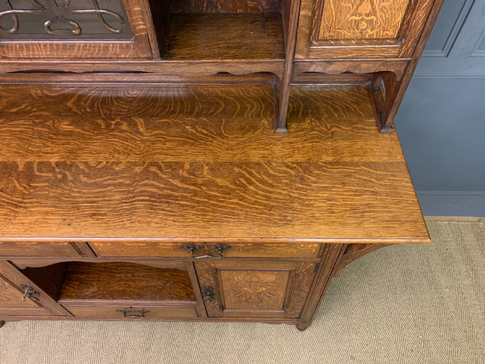An Arts and Crafts oak sideboard featuring detailed inlaid floral marquetry panels, a central leaded glass upper cabinet, and decorative copper hardware. View XVII