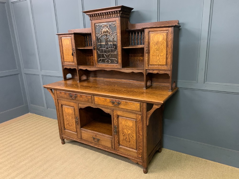 An Arts and Crafts oak sideboard featuring detailed inlaid floral marquetry panels, a central leaded glass upper cabinet, and decorative copper hardware. View XIX
