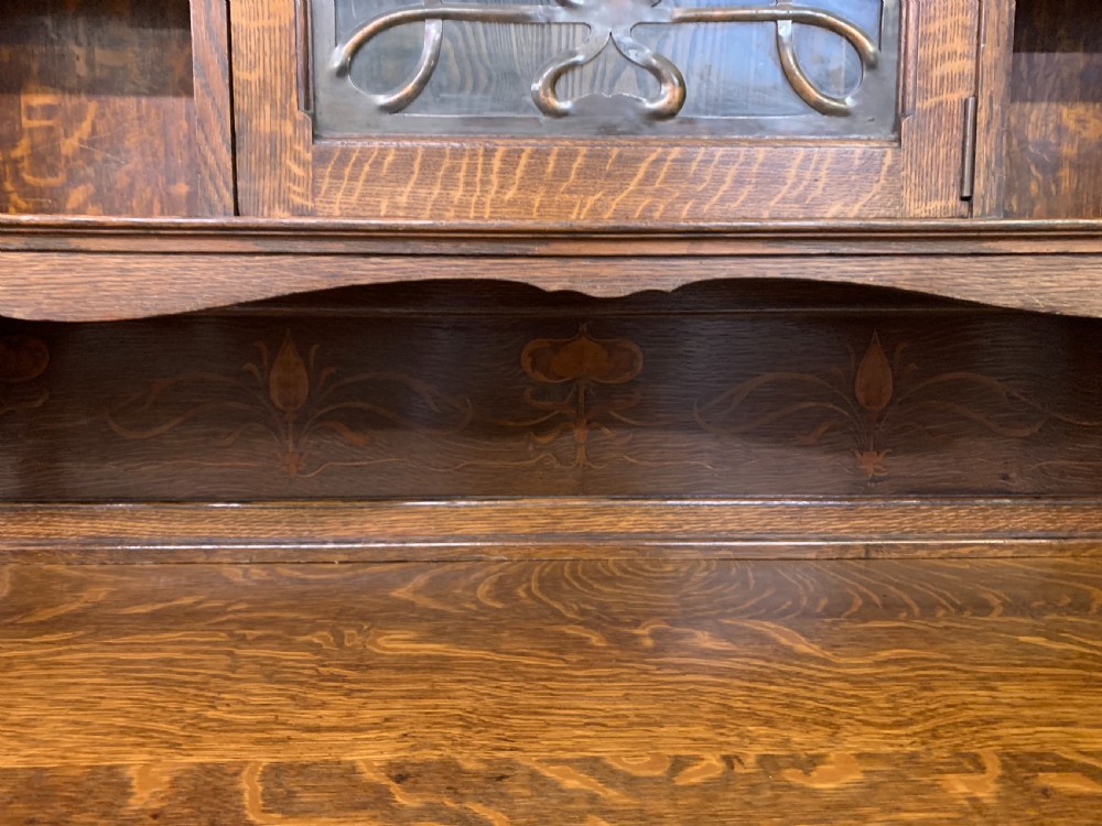 An Arts and Crafts oak sideboard featuring detailed inlaid floral marquetry panels, a central leaded glass upper cabinet, and decorative copper hardware. View XXII