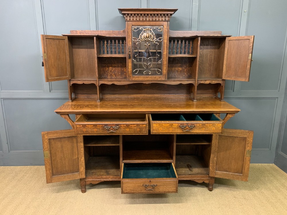 An Arts and Crafts oak sideboard featuring detailed inlaid floral marquetry panels, a central leaded glass upper cabinet, and decorative copper hardware. View XXV