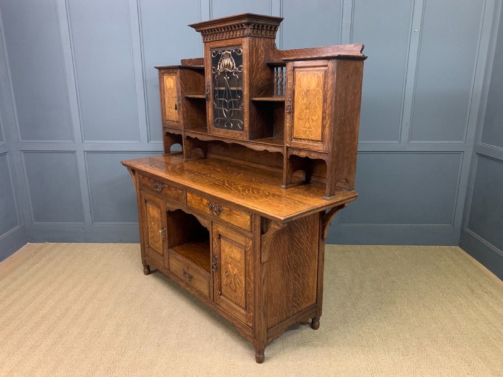 An Arts and Crafts oak sideboard featuring detailed inlaid floral marquetry panels, a central leaded glass upper cabinet, and decorative copper hardware. View XXVI