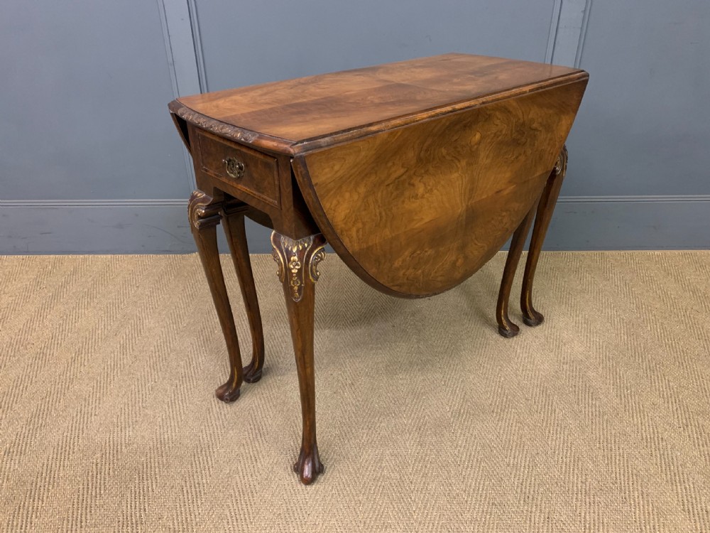 A burr walnut drop flap table featuring carved cabriole legs with leaf decoration, a single drawer with brass pull, and two rounded drop leaves.