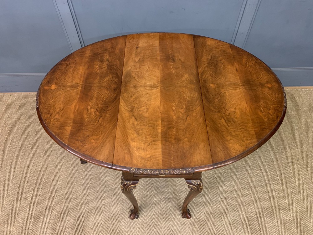 A burr walnut drop flap table featuring carved cabriole legs with leaf decoration, a single drawer with brass pull, and two rounded drop leaves. View VI