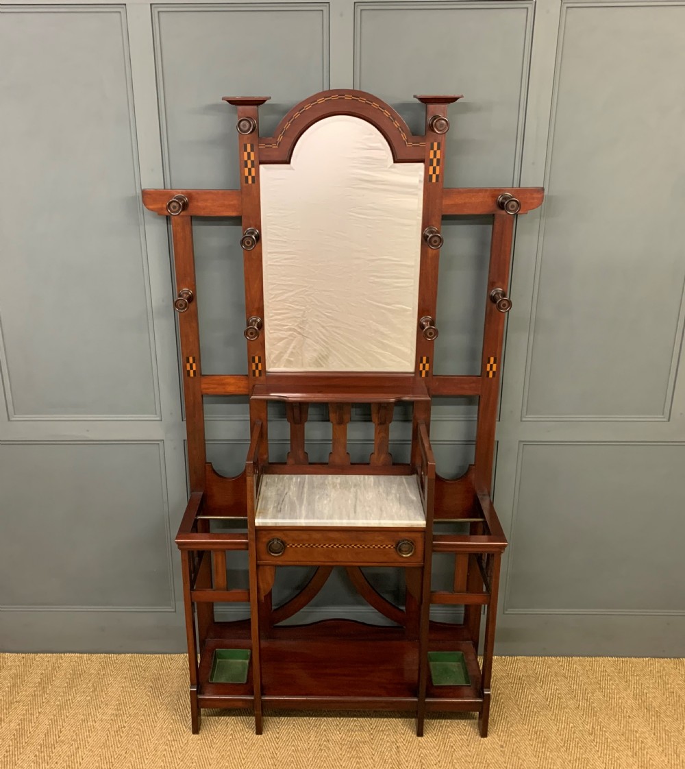 An Arts and Crafts inlaid mahogany hall stand featuring an arched central mirror, coat pegs, marble surface, and integrated umbrella racks.