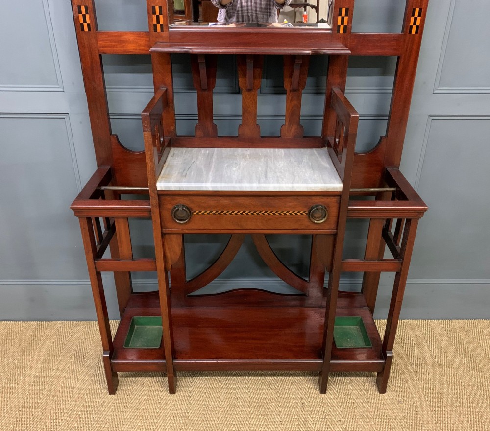 An Arts and Crafts inlaid mahogany hall stand featuring an arched central mirror, coat pegs, marble surface, and integrated umbrella racks. View III