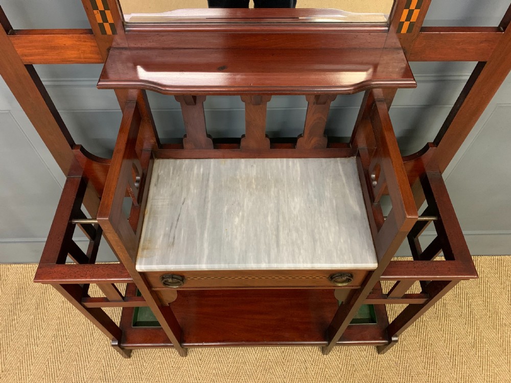 An Arts and Crafts inlaid mahogany hall stand featuring an arched central mirror, coat pegs, marble surface, and integrated umbrella racks. View VI