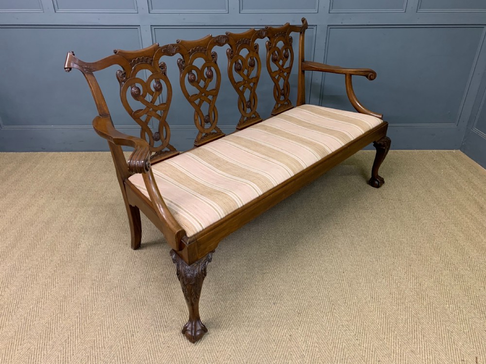 A carved mahogany Chippendale style settee featuring four pierced splats, scrolled arms, a striped fabric seat, and ball-and-claw feet.