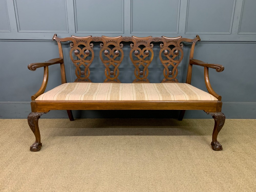 A carved mahogany Chippendale style settee featuring four pierced splats, scrolled arms, a striped fabric seat, and ball-and-claw feet. View V