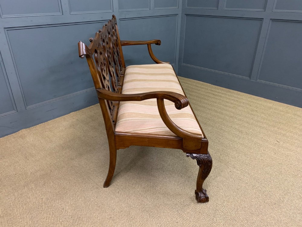 A carved mahogany Chippendale style settee featuring four pierced splats, scrolled arms, a striped fabric seat, and ball-and-claw feet. View XVII