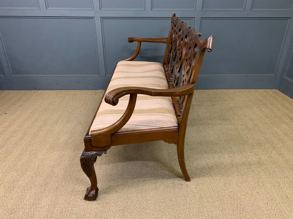 A carved mahogany Chippendale style settee featuring four pierced splats, scrolled arms, a striped fabric seat, and ball-and-claw feet. View XX