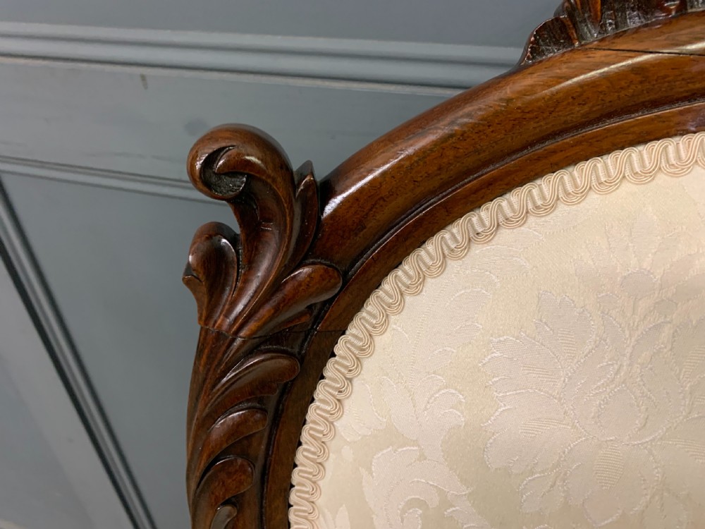 A Victorian rosewood high-back chair featuring intricately carved scrollwork, cream patterned damask upholstery, and cabriole front legs with brass castors. View V