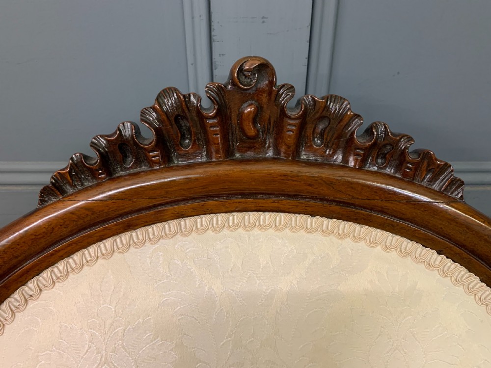 A Victorian rosewood high-back chair featuring intricately carved scrollwork, cream patterned damask upholstery, and cabriole front legs with brass castors. View VI