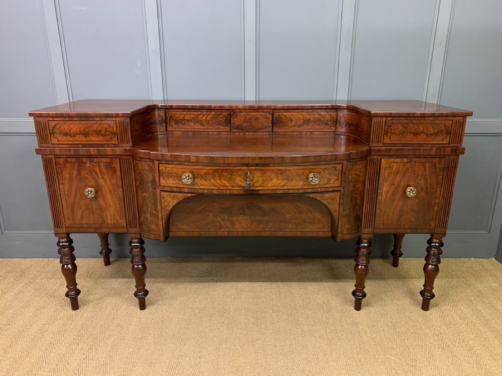 A Regency period flame mahogany sideboard featuring a raised stage top, central drawer, two side cupboards, detailed stringing, and turned reeded legs.